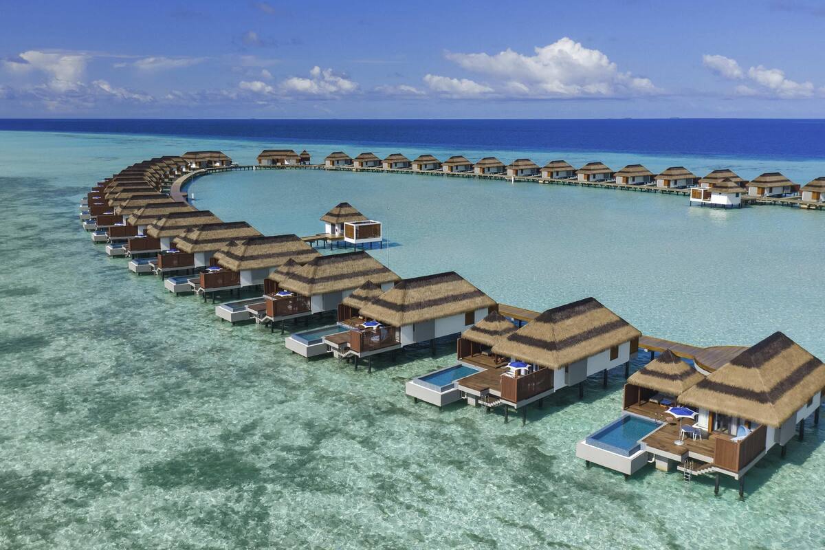 Cabañas sobre el agua dispuestas en forma de media luna, conectadas por pasarelas, con agua turquesa cristalina y un cielo despejado.