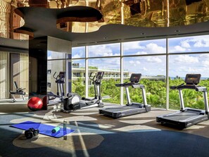 A gym with treadmills, exercise bikes, and a yoga mat area, with large windows offering a view of buildings and greenery.