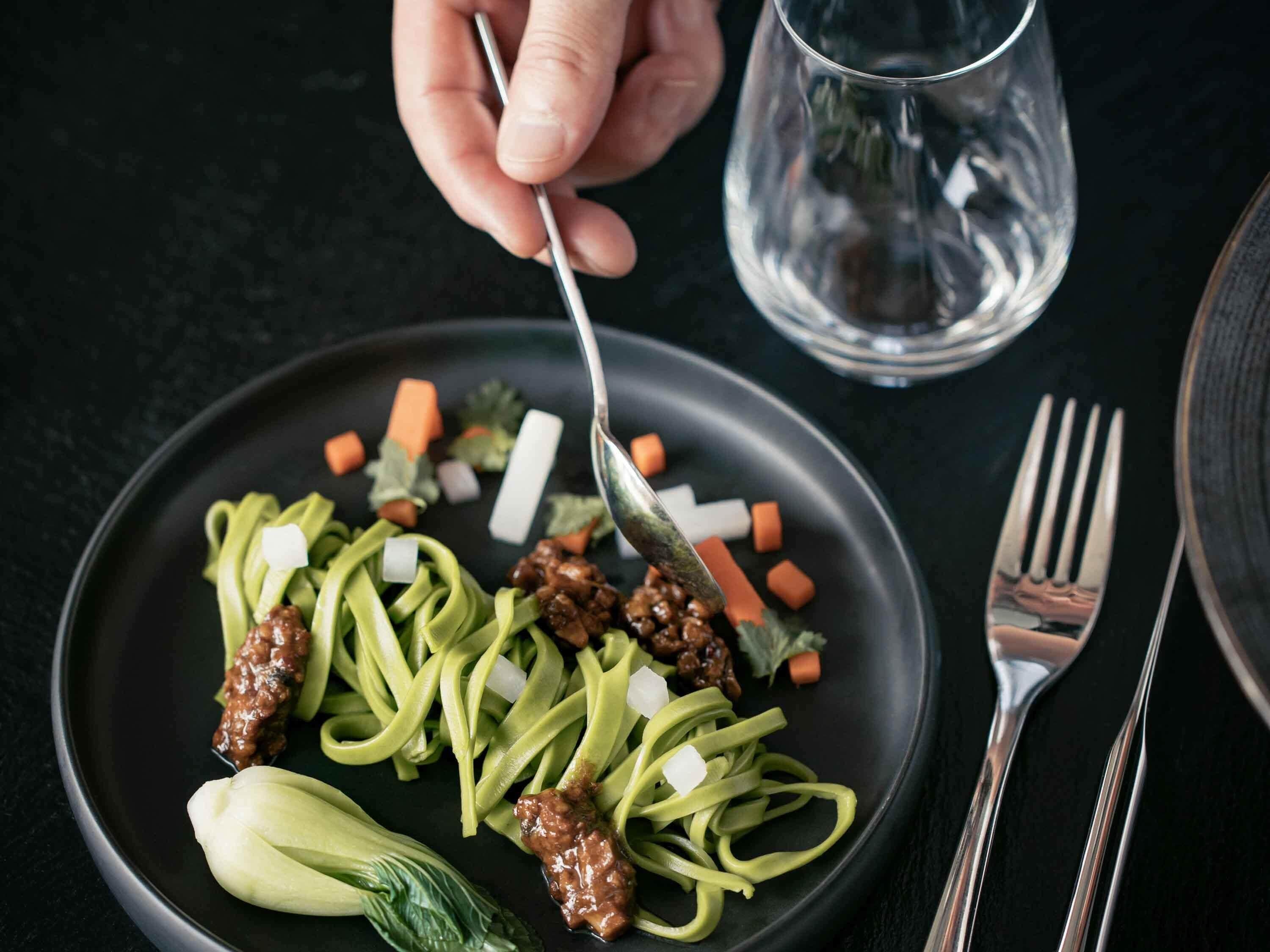A plate with noodles, vegetables, and meat, a fork, and a glass of water on a dark surface.