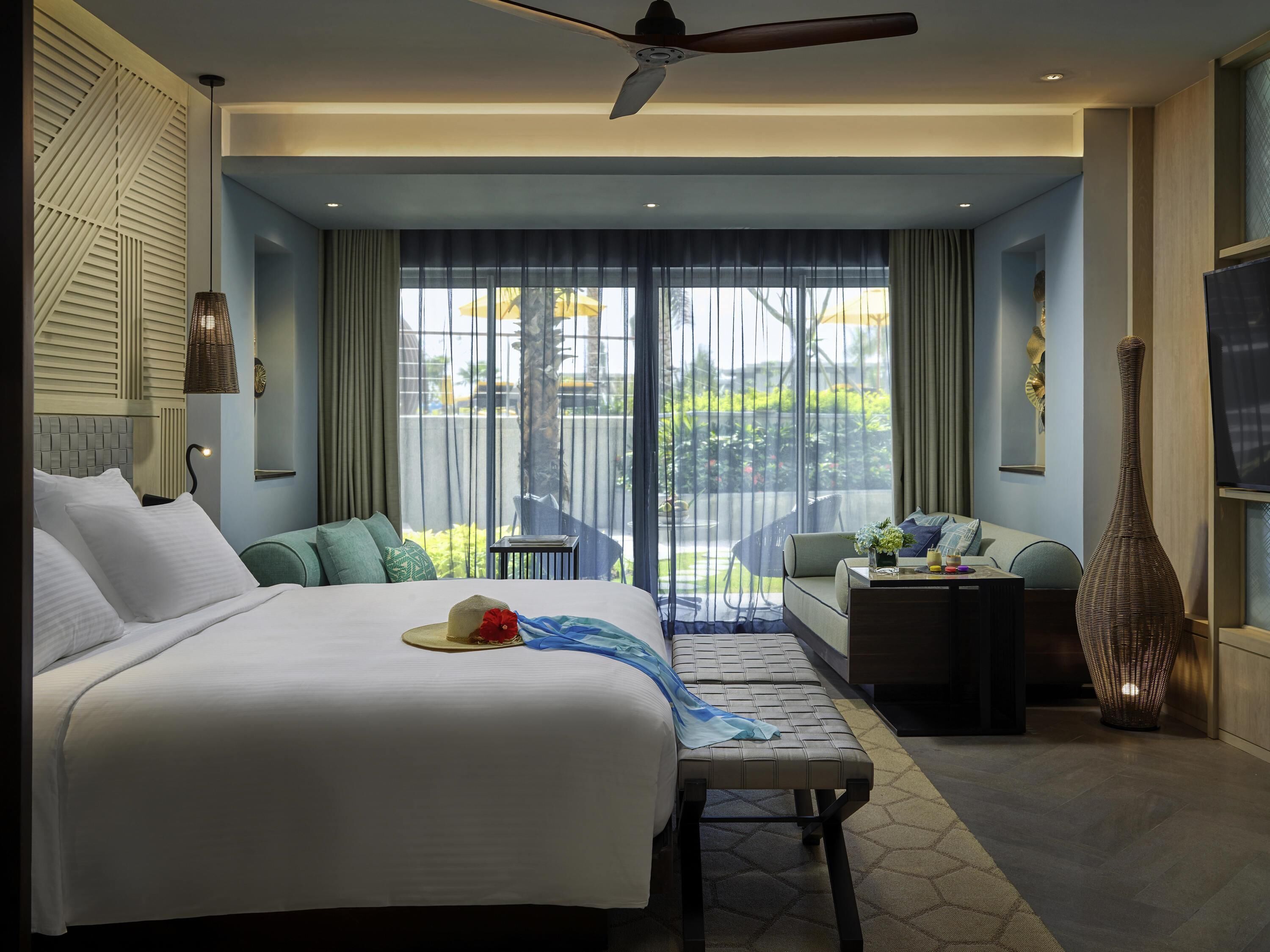 A modern hotel room with a large bed, a seating area, a TV, and a balcony with a view.