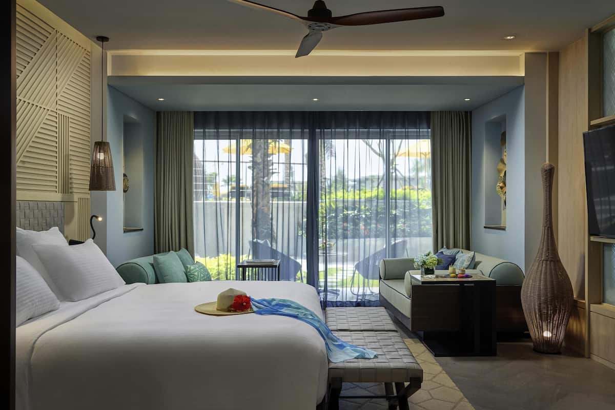 A modern hotel room with a large bed, a seating area, a TV, and a balcony with a view.