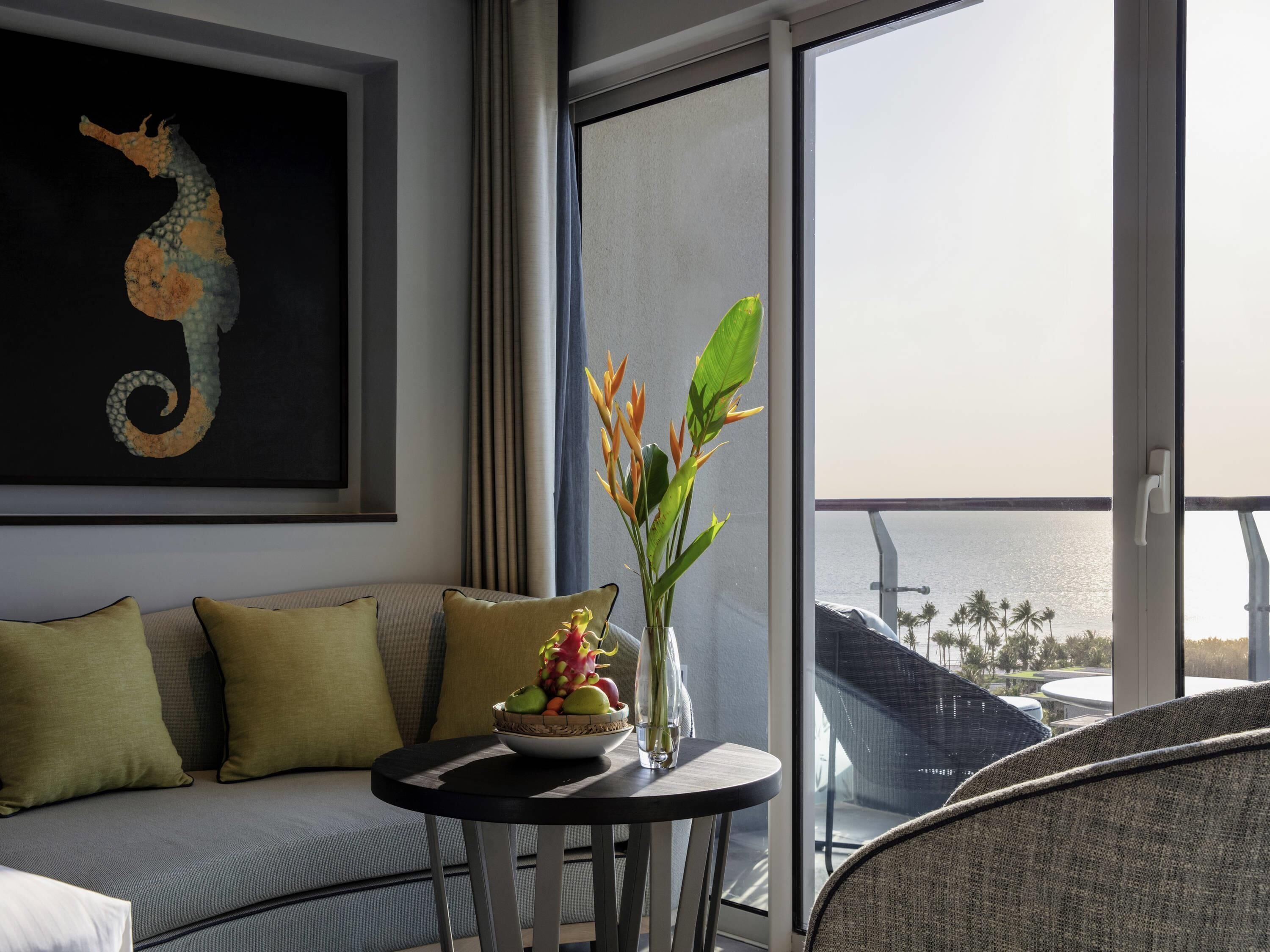 Een balkon met uitzicht op zee, een bank met kussens, een tafel met een schaal fruit en bloemen, en een schilderij van een zeepaard aan de muur.