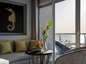 Varanda vista para o mar, sofá com almofadas, mesa com uma tigela de frutas e flores, e um quadro de um cavalinho-marinho na parede.