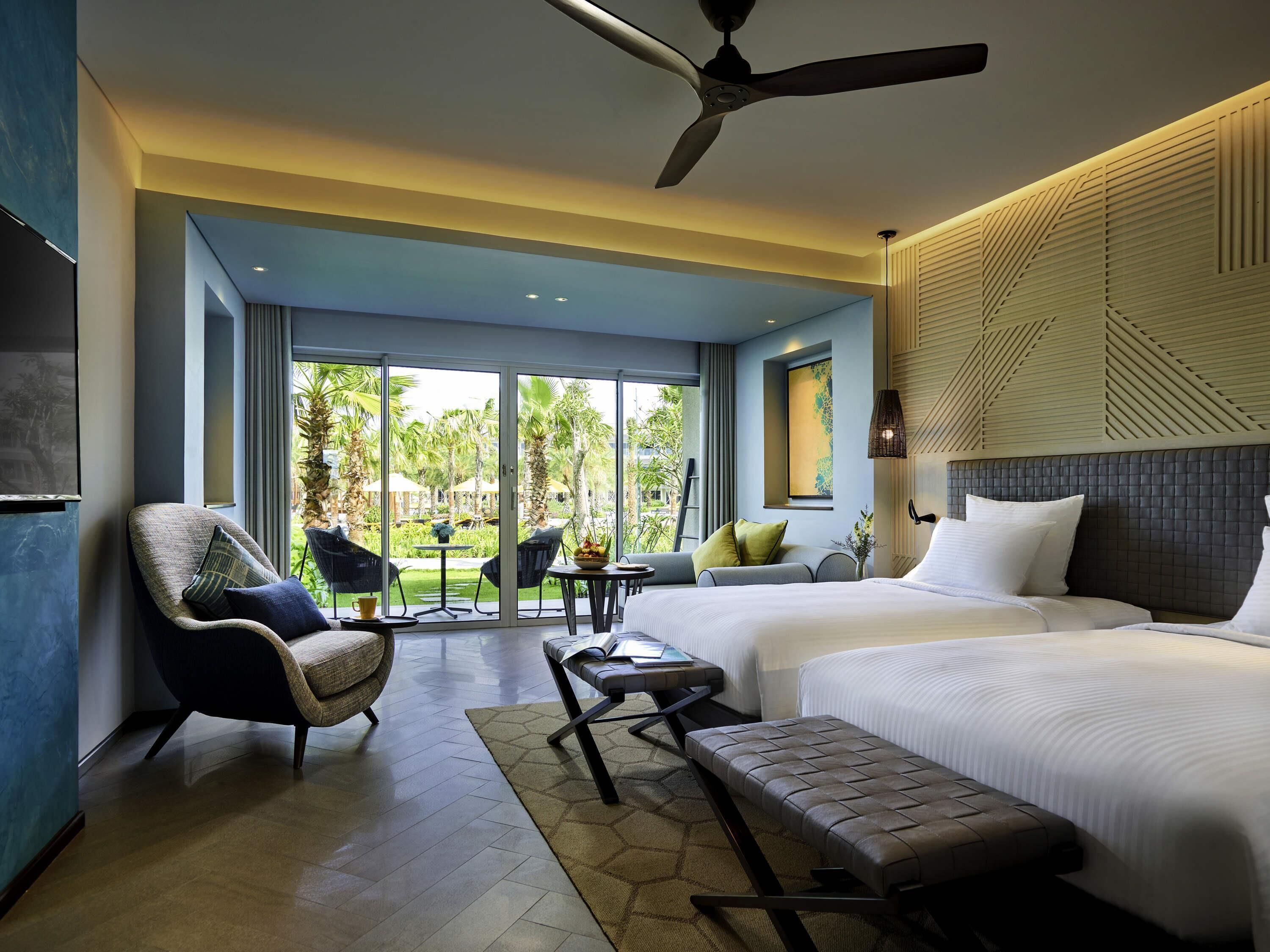 A modern hotel room with a large bed, a sofa, a chair, a small table, and a ceiling fan.