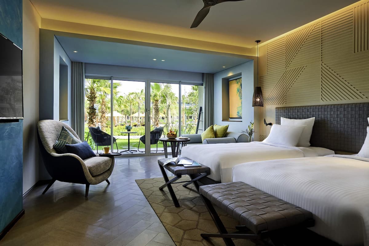 A modern hotel room with a large bed, a sofa, a chair, a small table, and a ceiling fan.