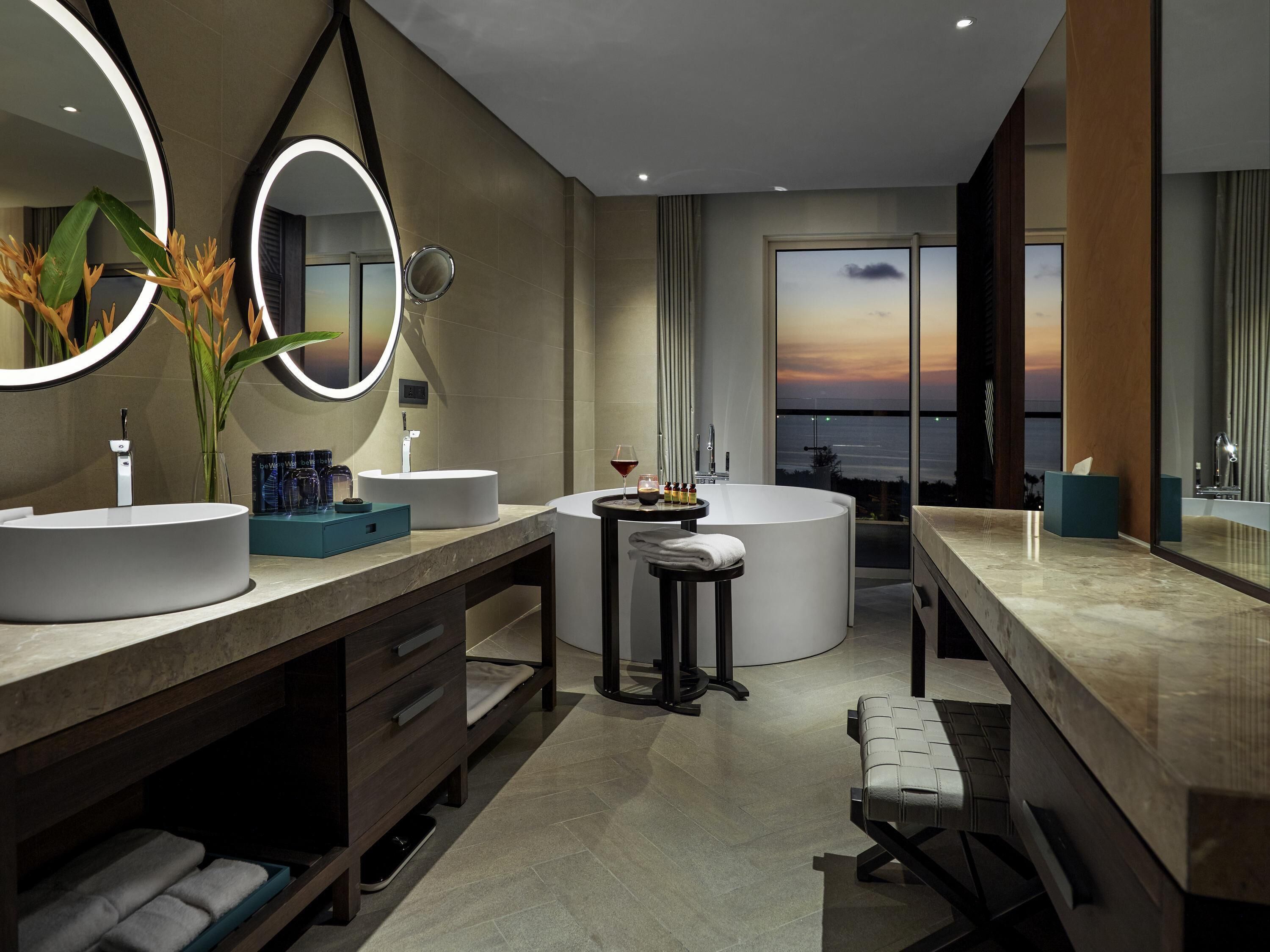 A modern bathroom with two sinks, a vanity, a round bathtub, and a large mirror with a light.