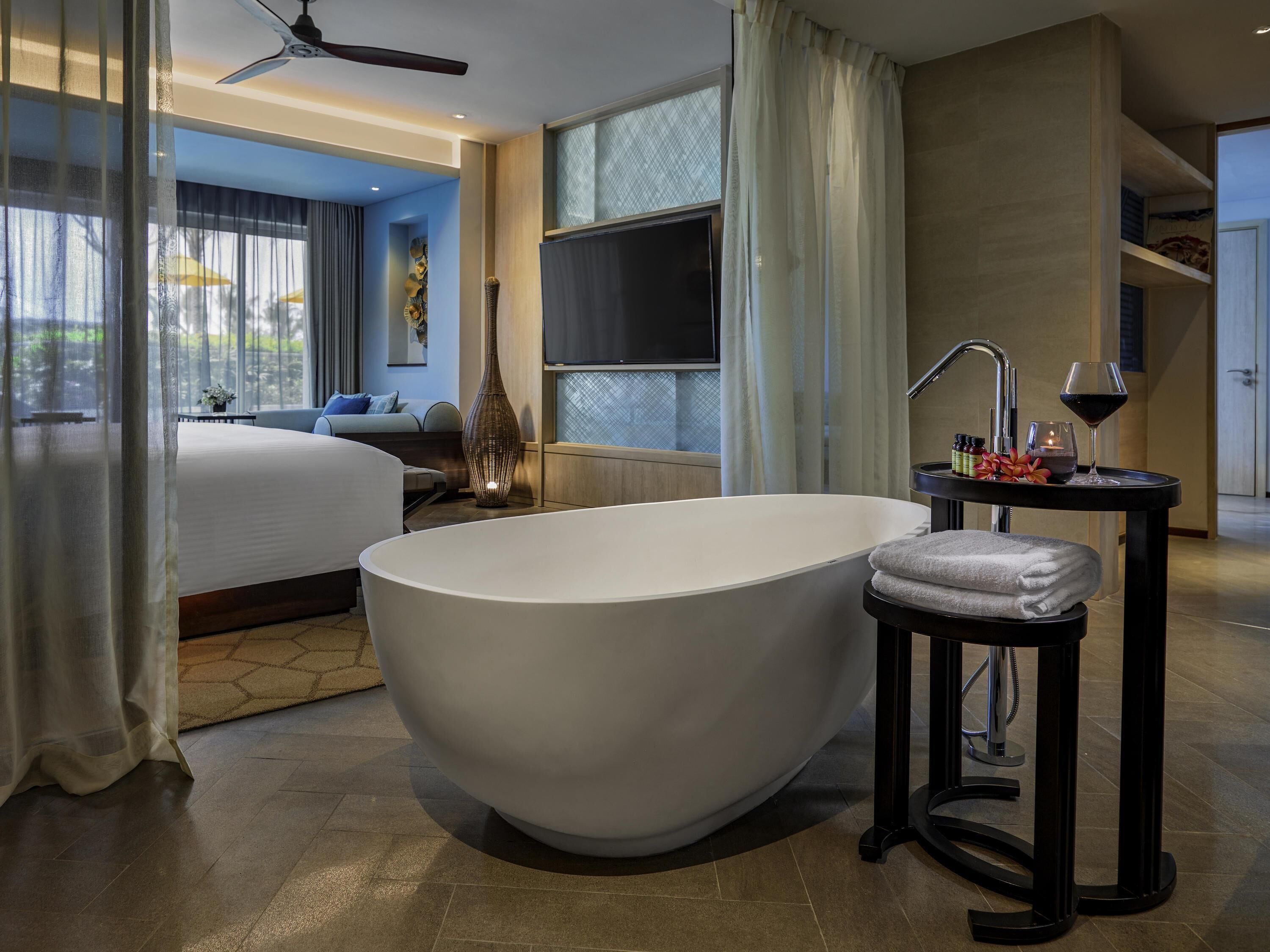 A modern hotel room with a large bathtub, a bed, a TV, and a small table with a wine glass and candles.