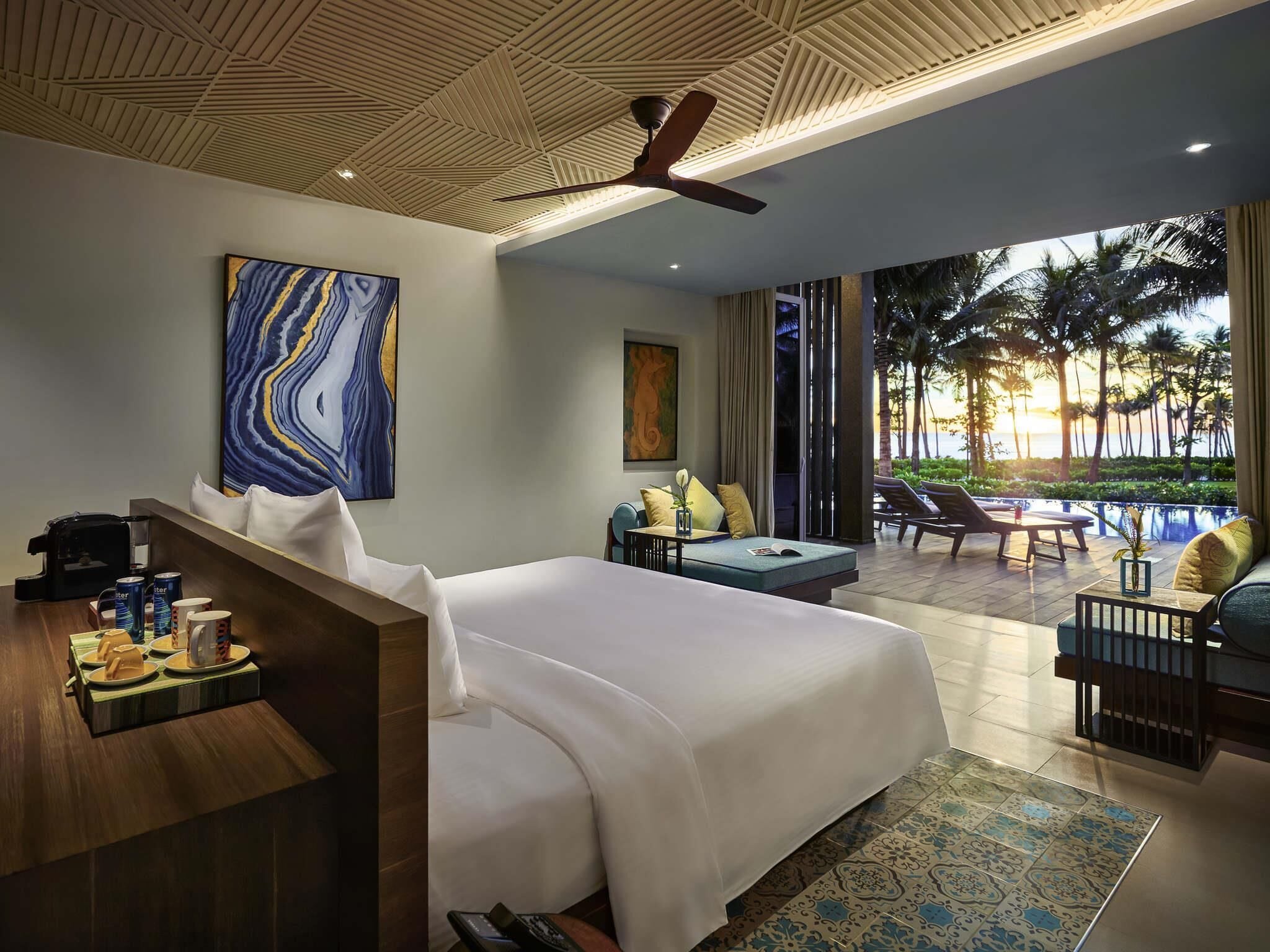 A hotel room with a bed, seating area, artwork, and a view of palm trees and the ocean.