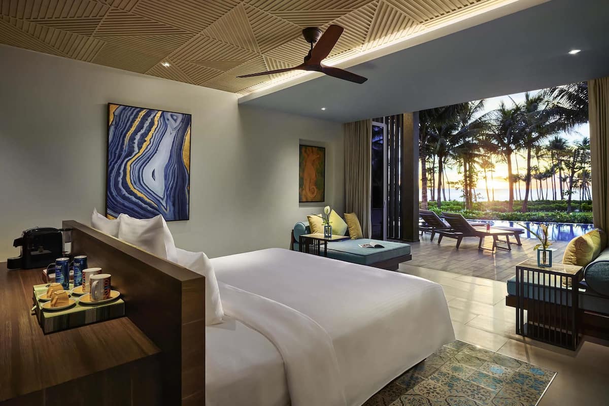 A hotel room with a bed, seating area, artwork, and a view of palm trees and the ocean.