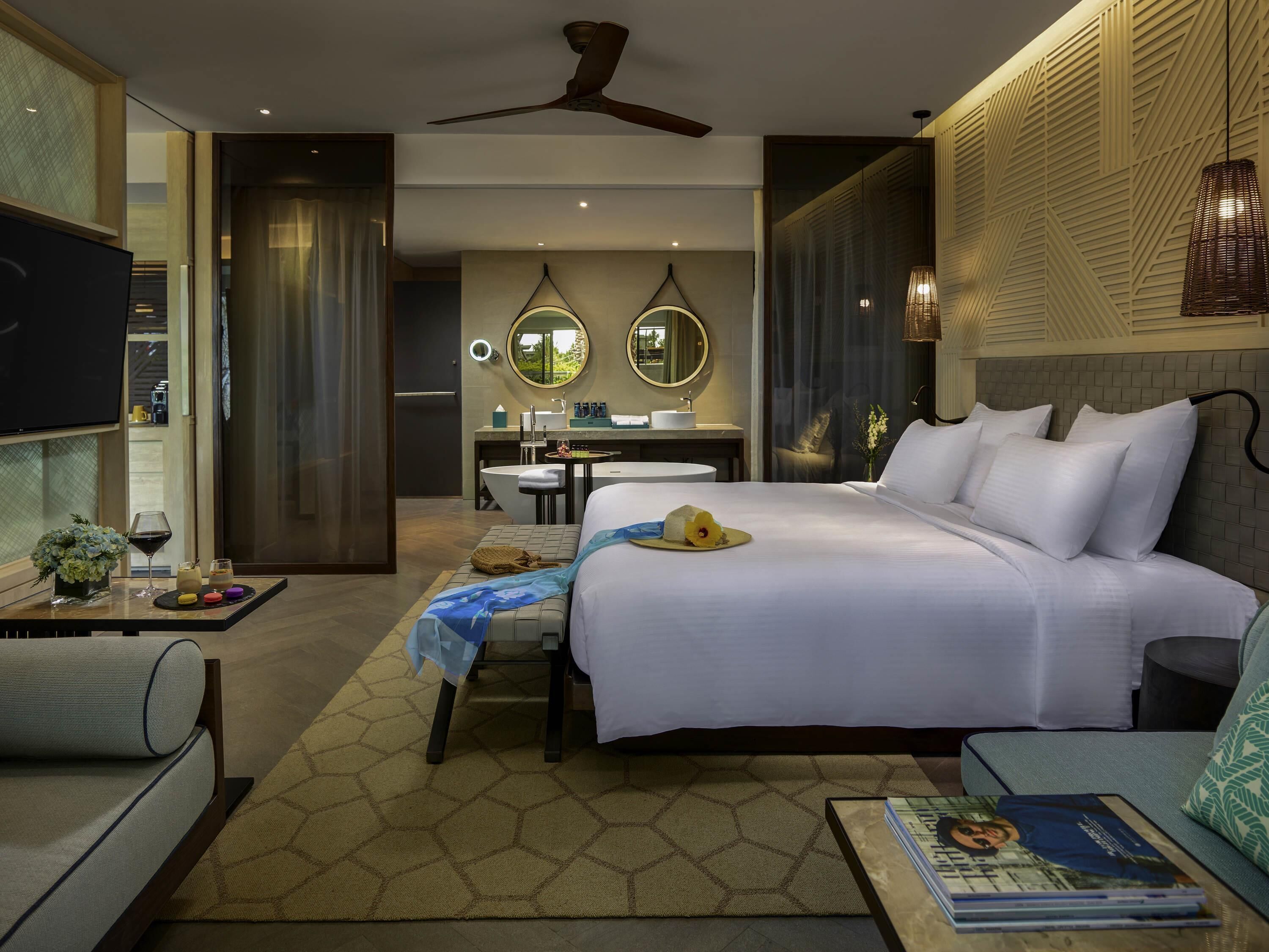 A hotel room with a bed, a desk, a chair, a TV, and a dining table with a plant and a wine glass.