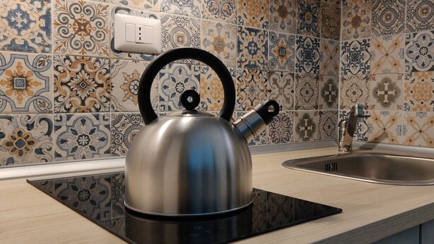 Una tetera de acero inoxidable sobre una cocina por inducción negra en una cocina con salpicadero de azulejos y un grifo moderno.