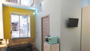 Habitación compacta con pared amarilla, ventana con rejas de metal, refrigerador blanco pequeño, microondas verde azulado, banca blanca y mesa blanca.