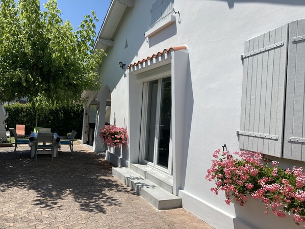 A white building with a tiled roof, a window with shutters, and a door. There are steps leading to the entrance, and a tree is visible to the left.