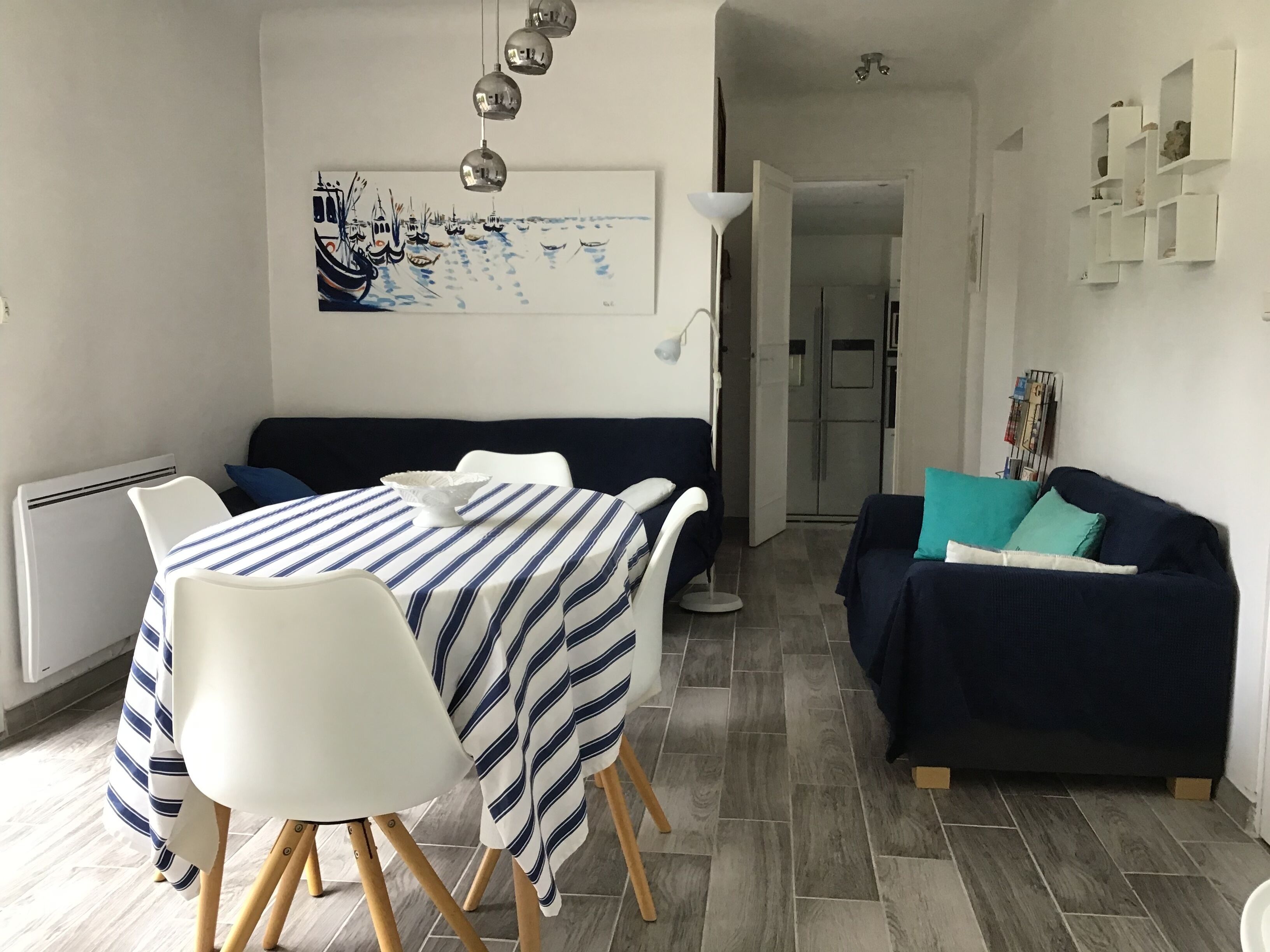 A modern living room with a dining area, featuring a striped tablecloth, white chairs, a dark blue sofa, and a painting on the wall.