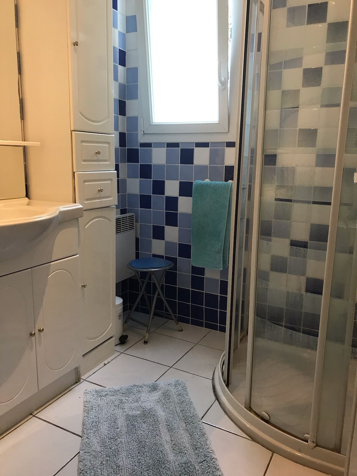 A bathroom with a white sink, a blue towel, a small stool, and a shower with a checkered glass door.