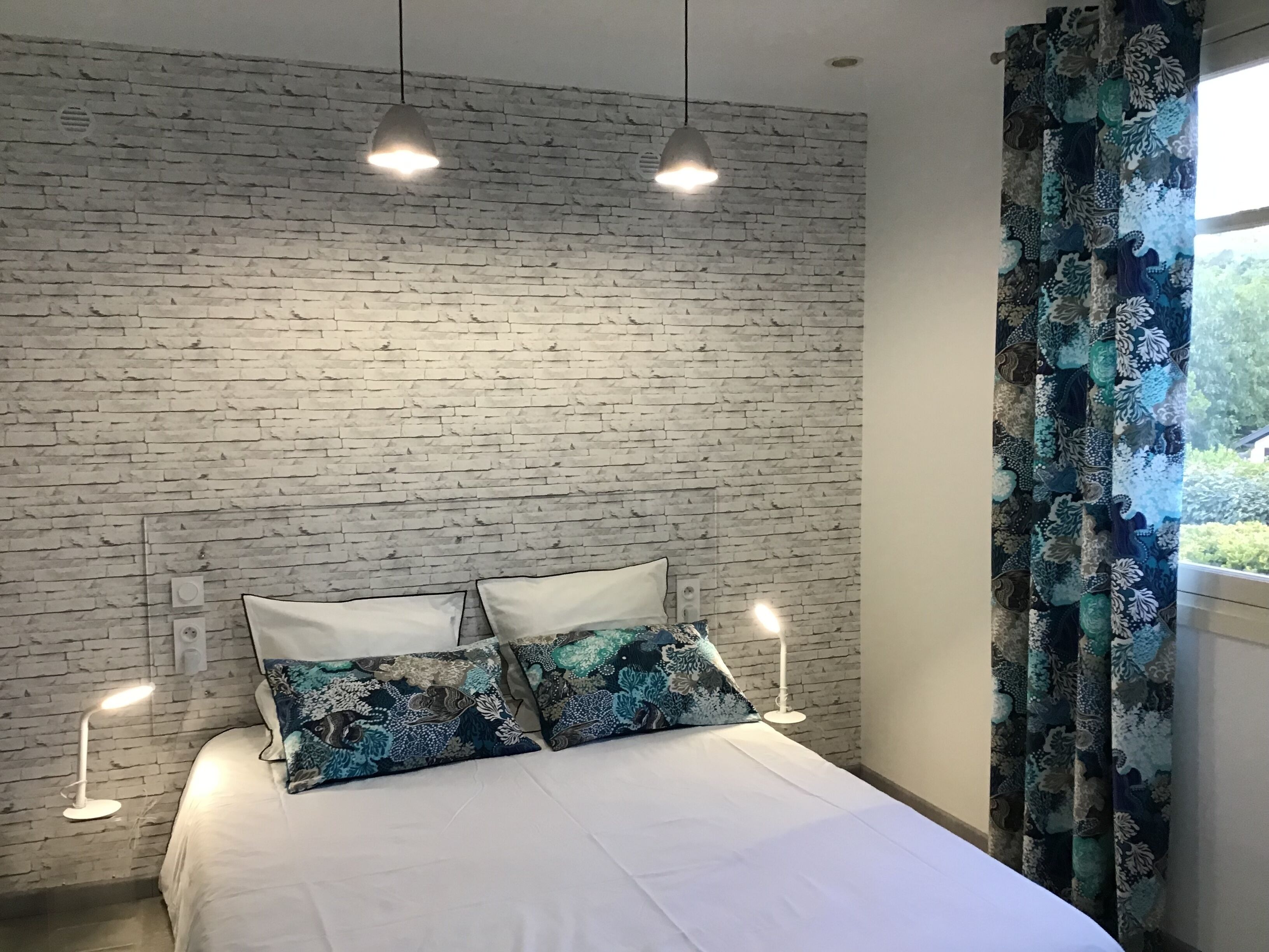A bedroom with a brick-patterned wall, a bed with white bedding, patterned pillows, and two bedside lamps.