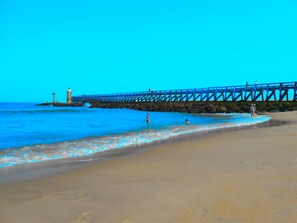 A beach with a pier extending into the water, featuring a lighthouse and rocky outcrops.