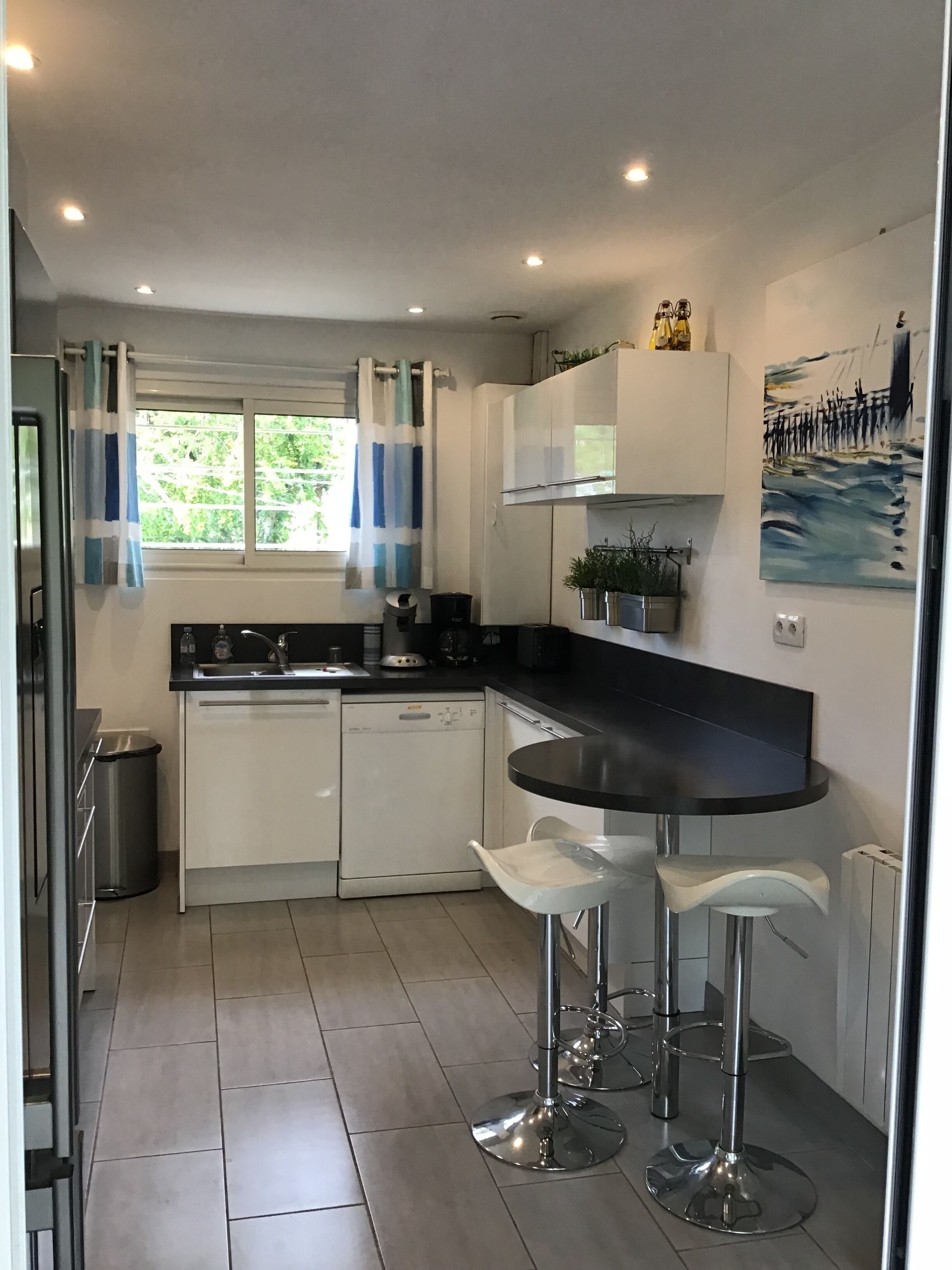 Een moderne keuken met witte kastjes, een zwart aanrecht en roestvrijstalen apparatuur. Er staan twee barkrukken bij en er is een raam met gordijnen.