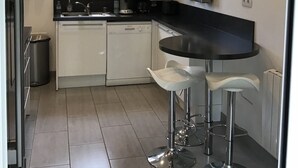 A modern kitchen with white cabinets, a black countertop, and stainless steel appliances. There are two bar stools and a window with curtains. - Nice Capbretonnaise three rooms between port, trade, and beach (Capbreton)