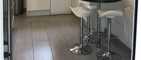 Une cuisine moderne avec des meubles blancs, un plan de travail noir et des appareils en acier inoxydable. On y trouve deux tabourets de bar et une fenêtre avec des rideaux.