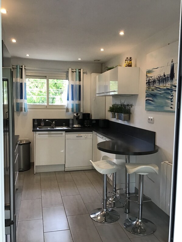 A modern kitchen with white cabinets, a black countertop, and stainless steel appliances. There are two bar stools and a window with curtains.