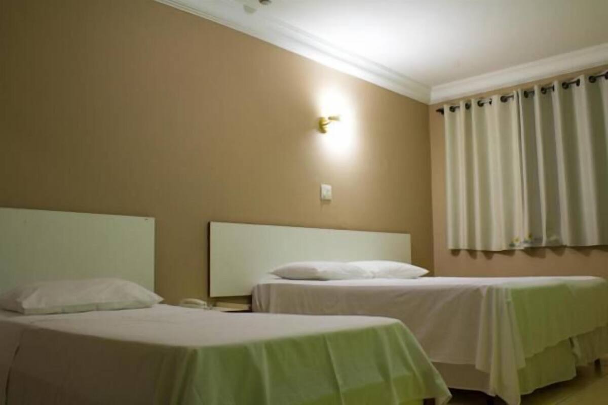 A hotel room with two single beds, a nightstand with a lamp, a wall-mounted light, and a window with curtains.