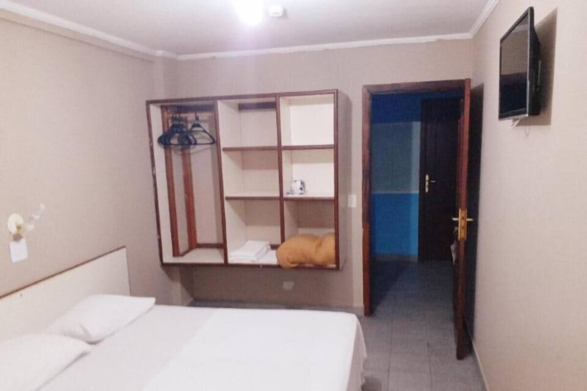 A hotel room with a bed, a wardrobe with shelves, a television, and a door leading to another room.