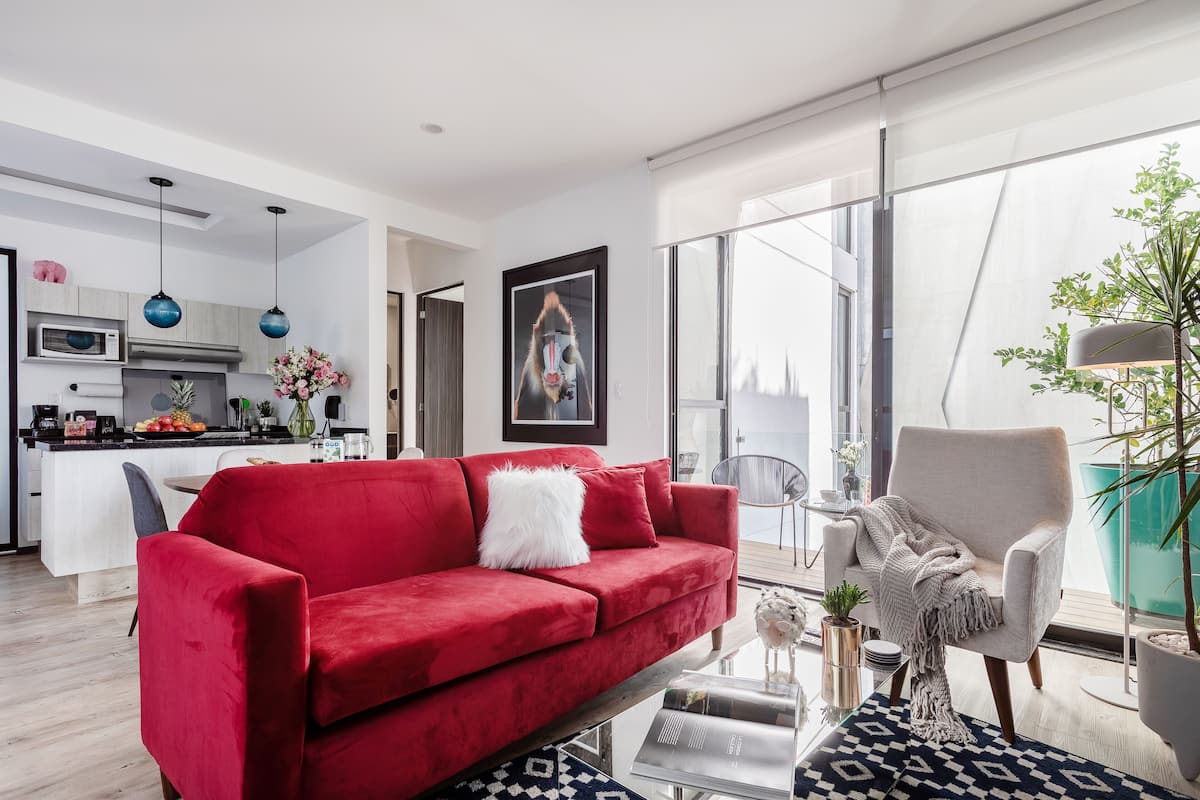 Ein modernes Wohnzimmer mit einem roten Sofa, einem weißen Sessel und einem Glastisch. Im Hintergrund ist eine Küche mit einer Mikrowelle und einer Obstschale zu sehen.