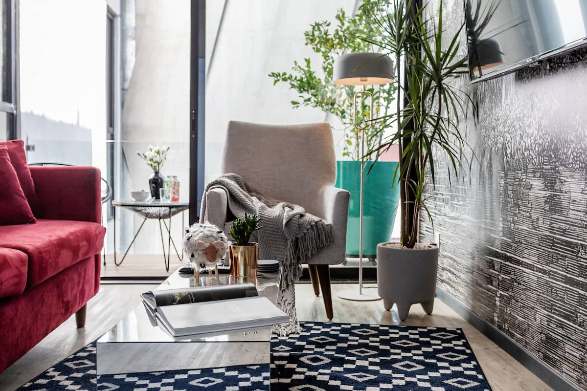 Ein modernes Wohnzimmer mit einem roten Sofa, einem grauen Sessel, einem Couchtisch mit Büchern, einer Stehlampe und einer großen Topfpflanze.