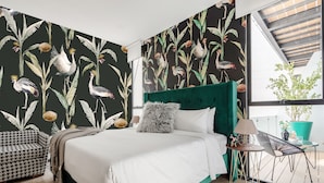 A bedroom with a large bed, a green upholstered headboard, a patterned wall, a small round table with a lamp, and a view of the outdoors. - Illumé Urban Living - Apartments in the heart of Roma Norte (Mexico City)
