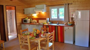 A compact kitchen with wooden walls, a dining table with chairs, a refrigerator, a microwave, a sink, and a window with curtains. - Accès Piscine, Fitness, Sauna ! Maisonnette Climatisée avec Terrasse (Gramat)
