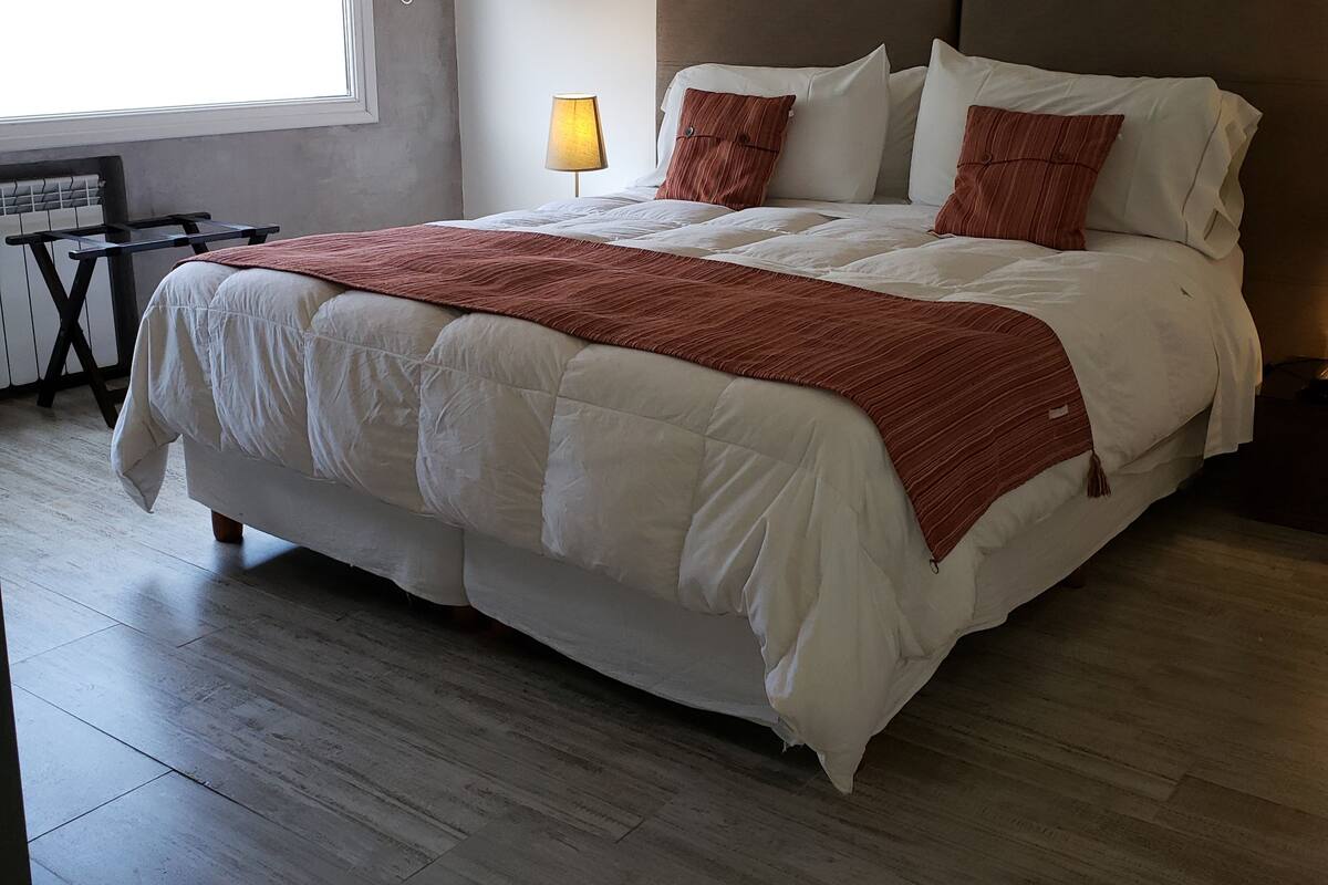 Habitación de hotel con una cama grande, una mesita de noche con lámpara, un cuadro enmarcado en la pared y una ventana con persianas.