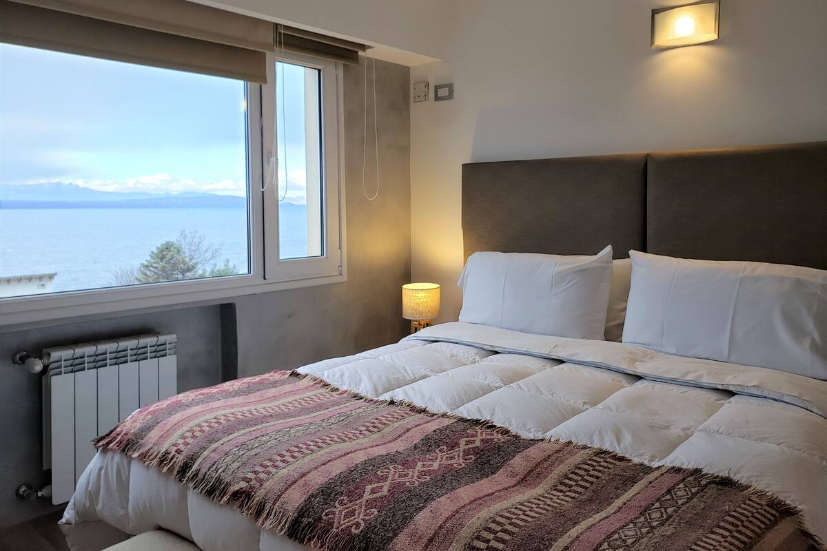Un dormitorio con cama, ventana con vista al mar, lámpara de pared y radiador.