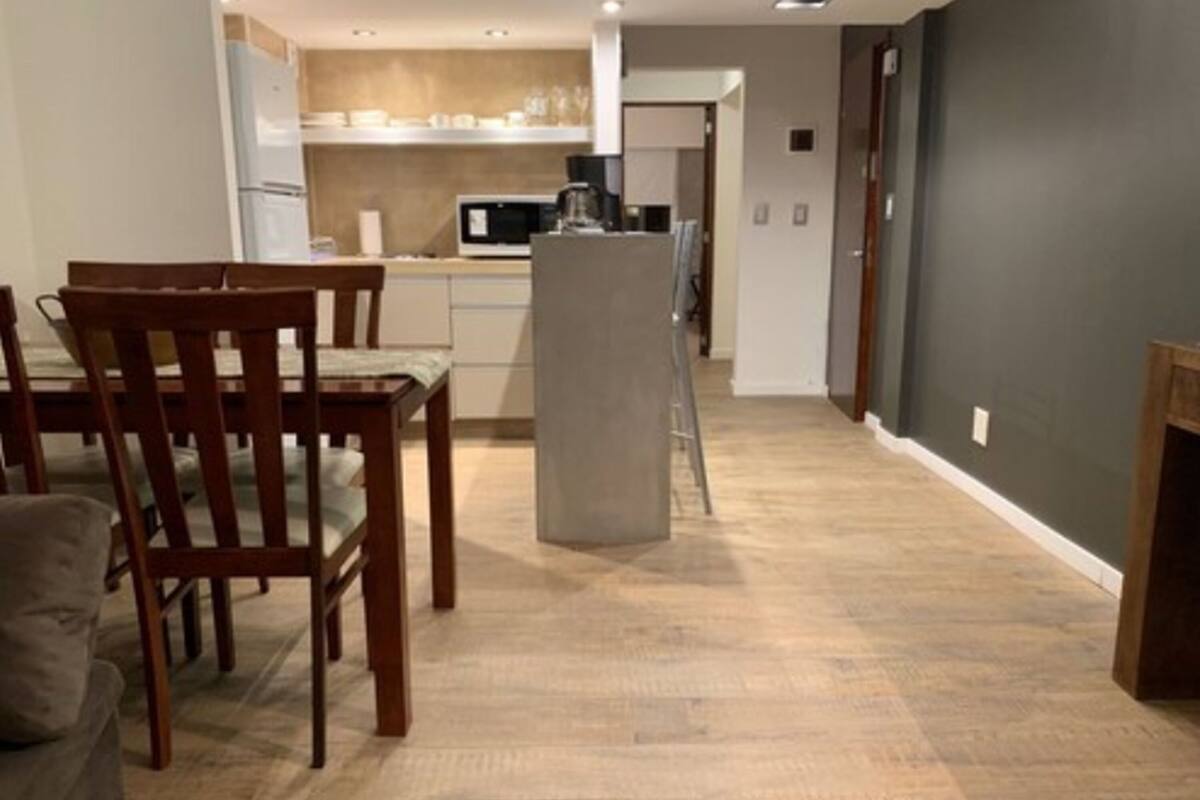 Una cocina moderna con armarios blancos, un microondas y una cafetera. Hay una zona de comedor con una mesa y sillas de madera.