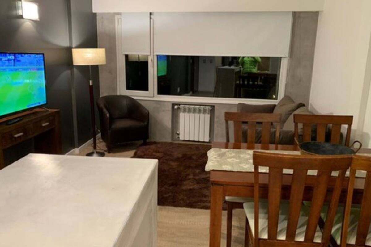 Una sala de estar con televisor, sofá, una mesa de comedor con sillas, una lámpara y una ventana con vista a la cocina.