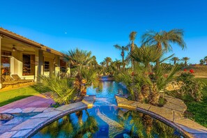 A pool area with a clear view of the surrounding landscape, including palm trees and a building with a tiled roof. - Spanish Palms - Lounge Poolside at PGA West! (La Quinta)