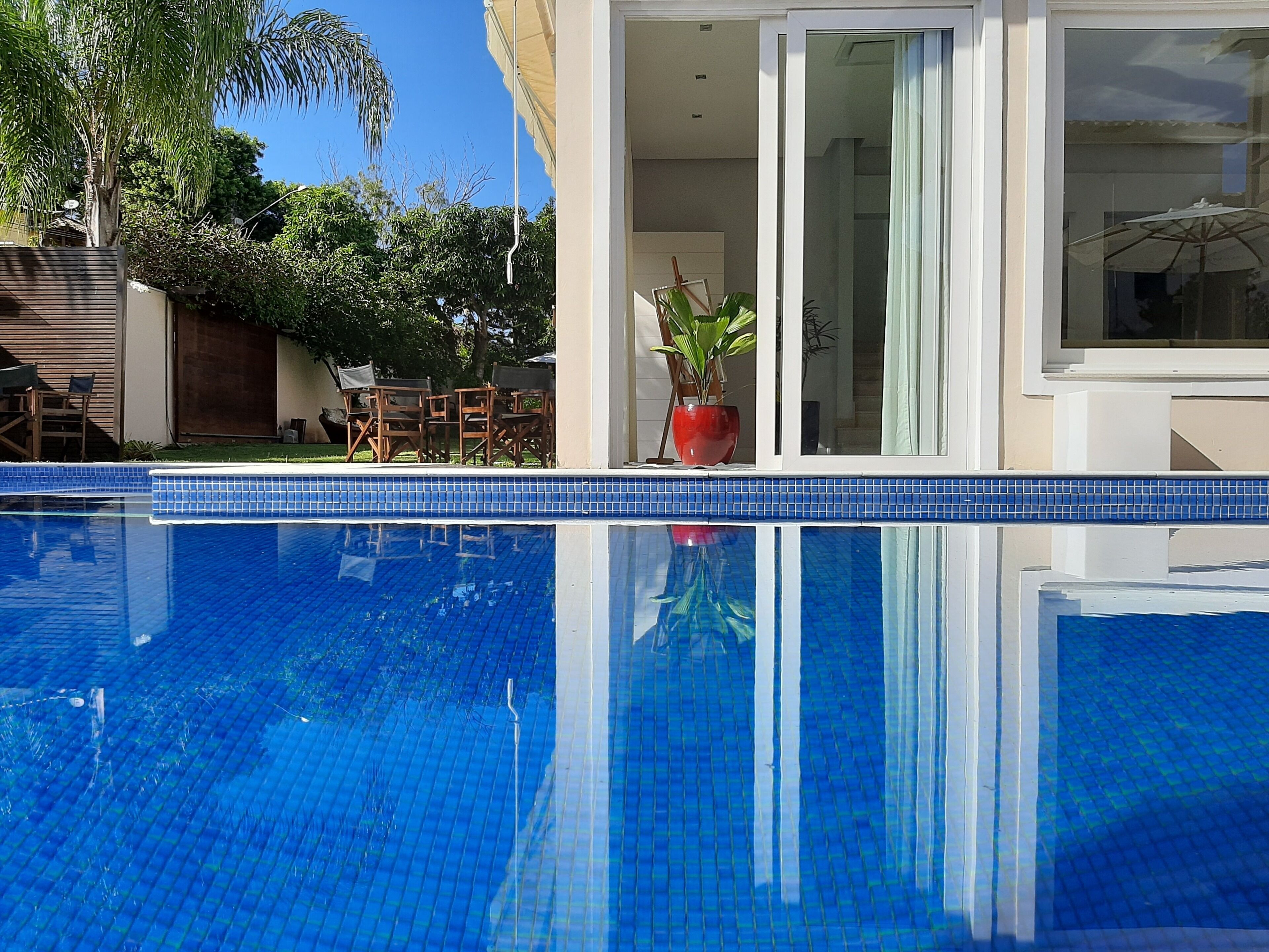A swimming pool with blue tiles, a patio area with wooden chairs, and a potted plant.