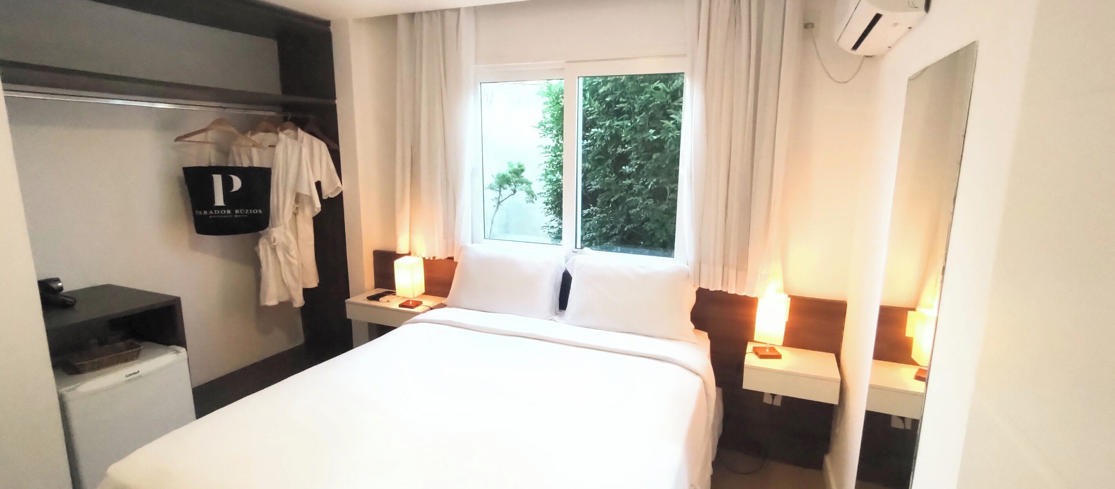 Una habitación de hotel con cama, mesitas de noche con lámparas, un armario, una ventana con vistas a zonas verdes y un equipo de aire acondicionado.