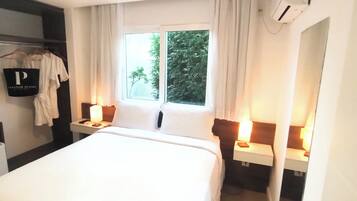 Una habitación de hotel con cama, mesitas de noche con lámparas, un armario, una ventana con vistas a zonas verdes y un equipo de aire acondicionado.