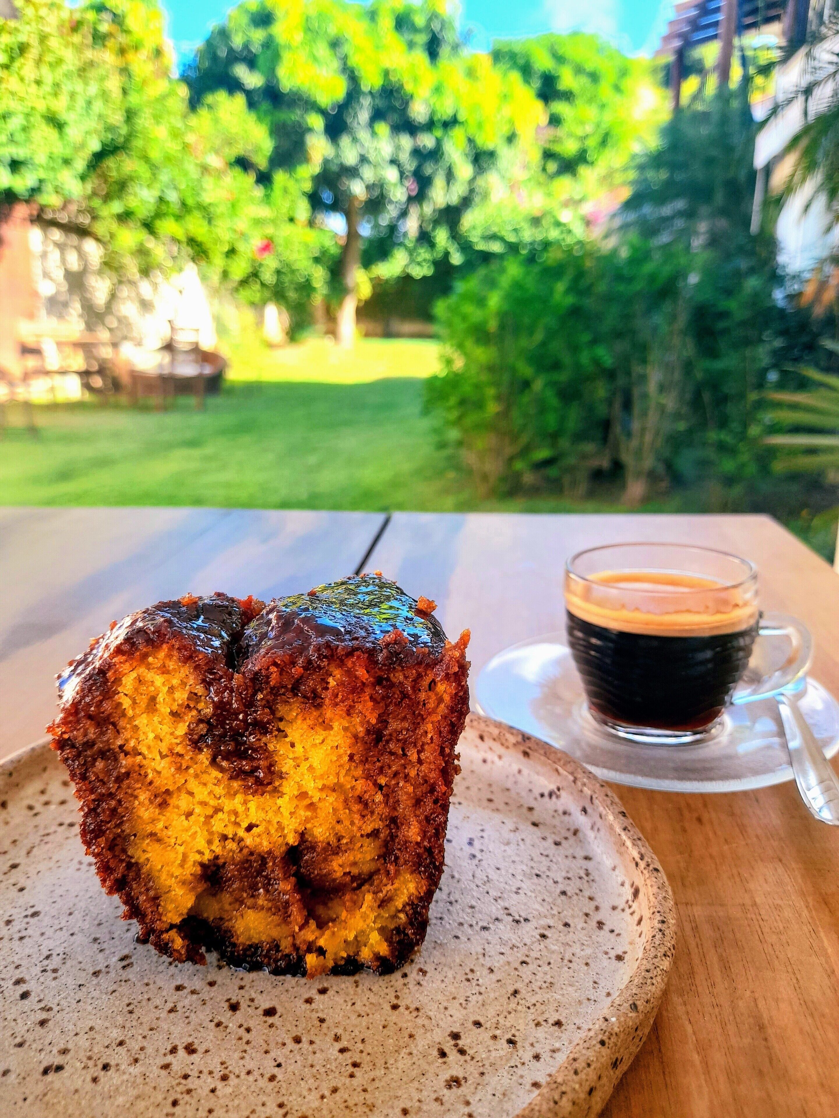 A slice of cake with a glossy chocolate topping on a ceramic plate, accompanied by a cup of dark coffee on a saucer.