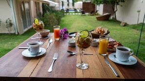 Ein Holztisch ist reichhaltig mit Frühstück gedeckt, darunter Obstsalat, Saft, Kaffee und Gebäck, mit Blick auf einen Garten und ein Gebäude im Hintergrund.