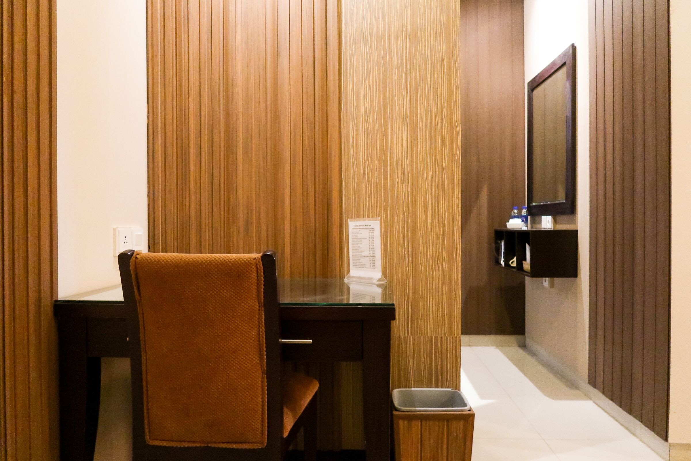 A hotel room with a wooden paneled wall, a glass desk, a chair with a towel, a trash bin, and a flat-screen TV mounted on the wall.