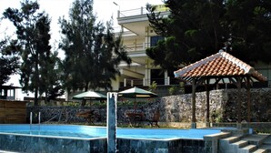 A swimming pool with a water feature, surrounded by lounge chairs and umbrellas, with a stone wall and trees in the background. - Tangko Inn Resort (Cipanas)