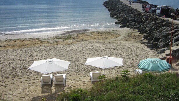 A beach with white lounge chairs and umbrellas, a rocky breakwater, and a calm sea. - Hotel Blian (Ravda)