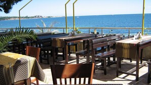 An outdoor dining area with wooden tables and benches, overlooking a body of water. - Hotel Blian (Ravda)