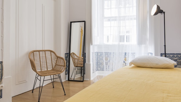 A bedroom with a bed, a chair, a mirror, and a window with sheer curtains.