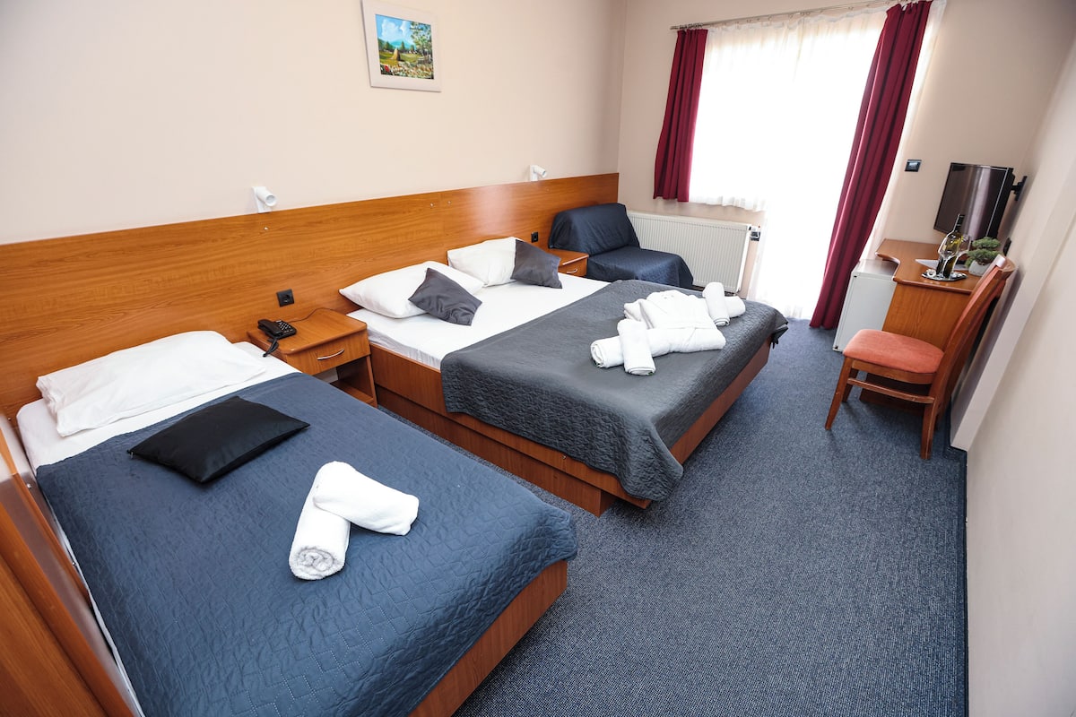 A hotel room with two single beds, a desk with a chair, a small refrigerator, and a window with curtains.