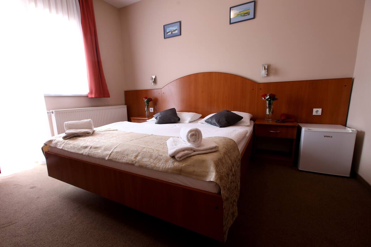A hotel room with a wooden bed, a nightstand, a small refrigerator, and a window with curtains.