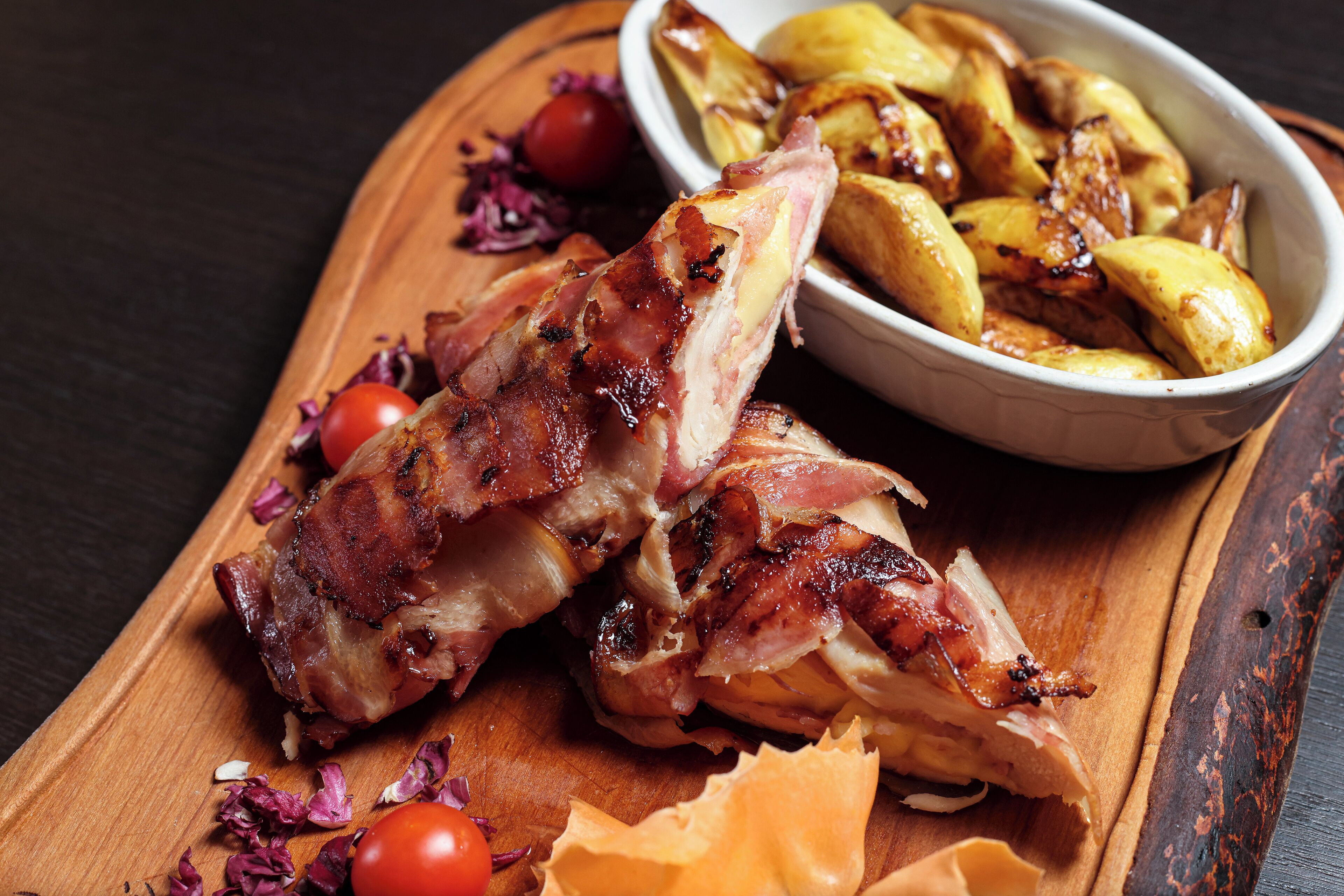 A wooden cutting board with roasted potatoes and a piece of meat, possibly pork, garnished with cherry tomatoes and red onion.
