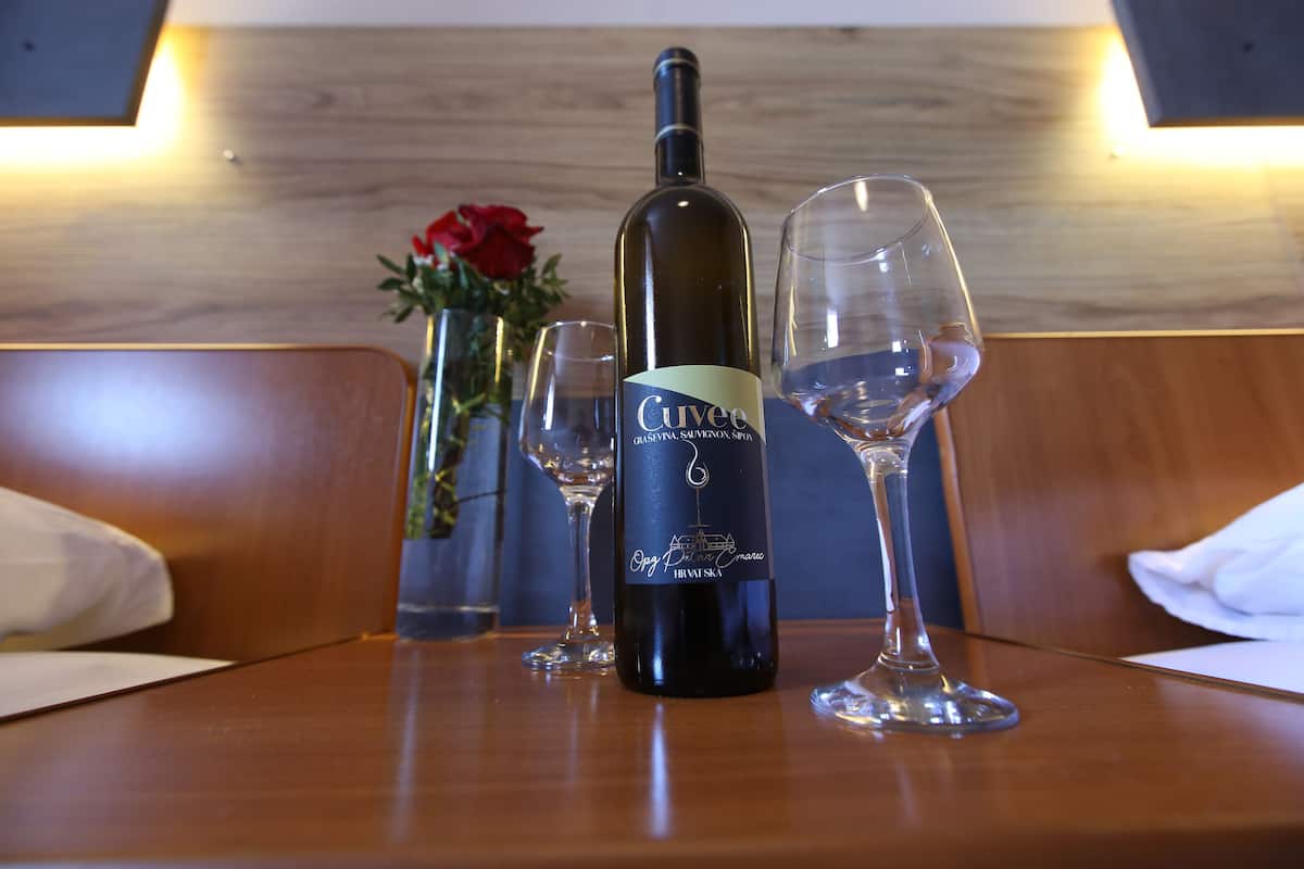 A bottle of Cuvee wine and two glasses on a wooden table in a room with a wooden wall and a vase with a red flower.